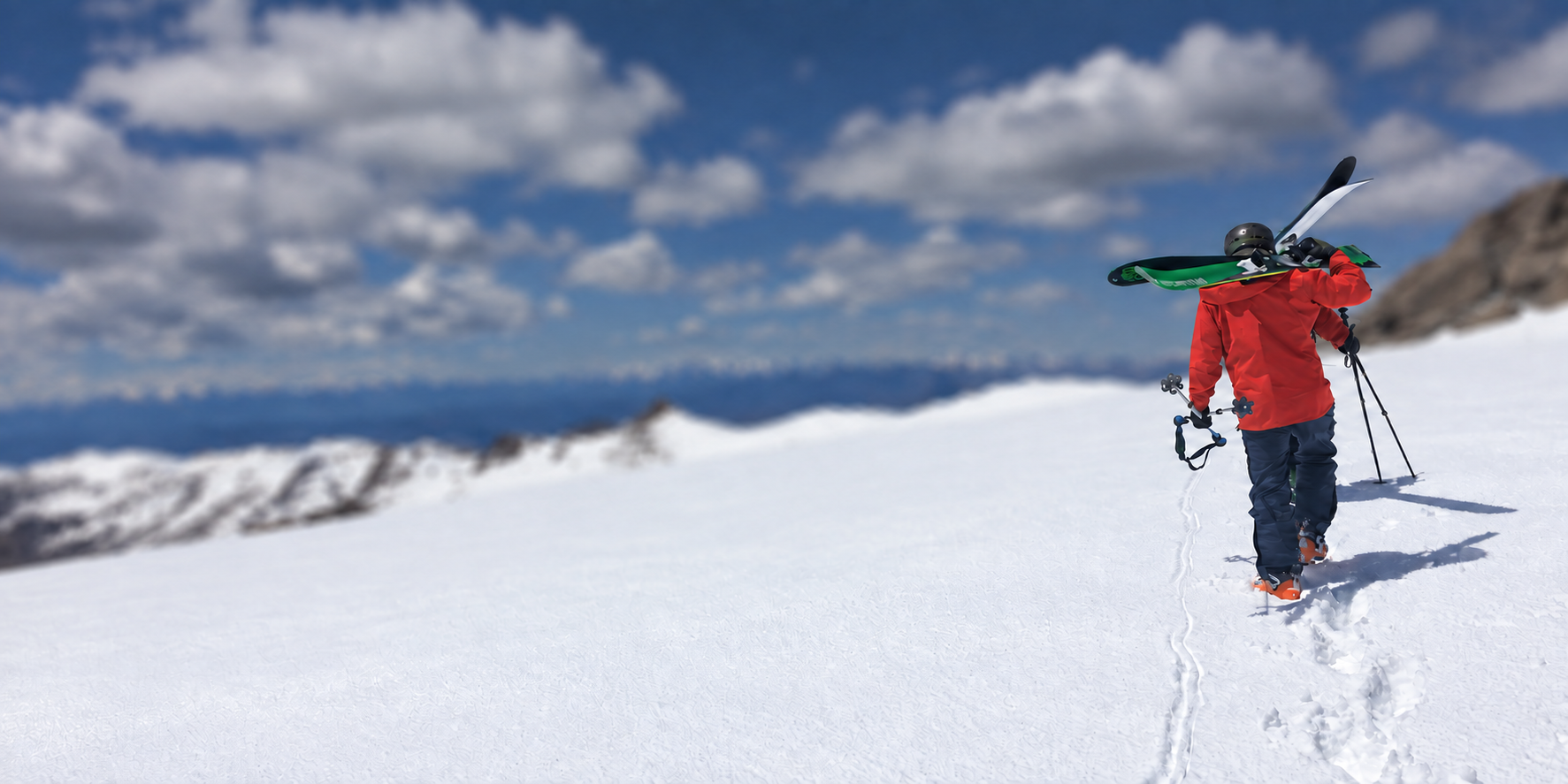 Skier in a red jacket walking uphill with skis, against softly blurred snowy mountains